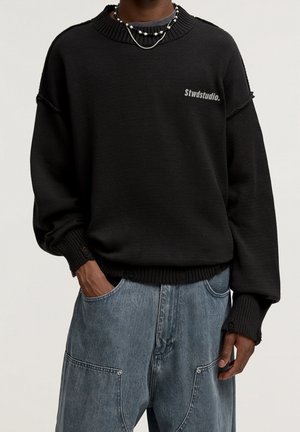 Person wearing a black Stwdstudio sweater with distressed cuffs and hem, paired with loose blue denim jeans and layered necklaces.