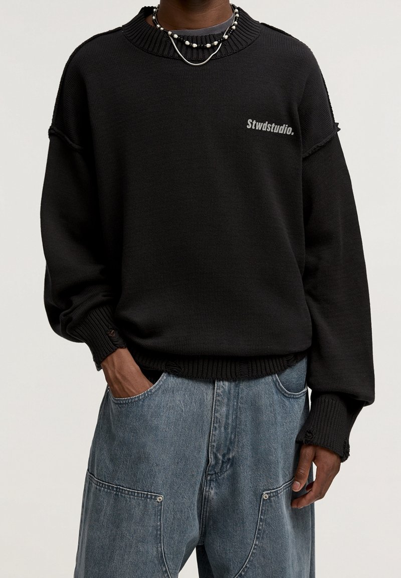 Personne portant un sweat-shirt noir Stwdstudio avec des poignets et un ourlet usés, associé à un jean en denim bleu ample et des colliers superposés.