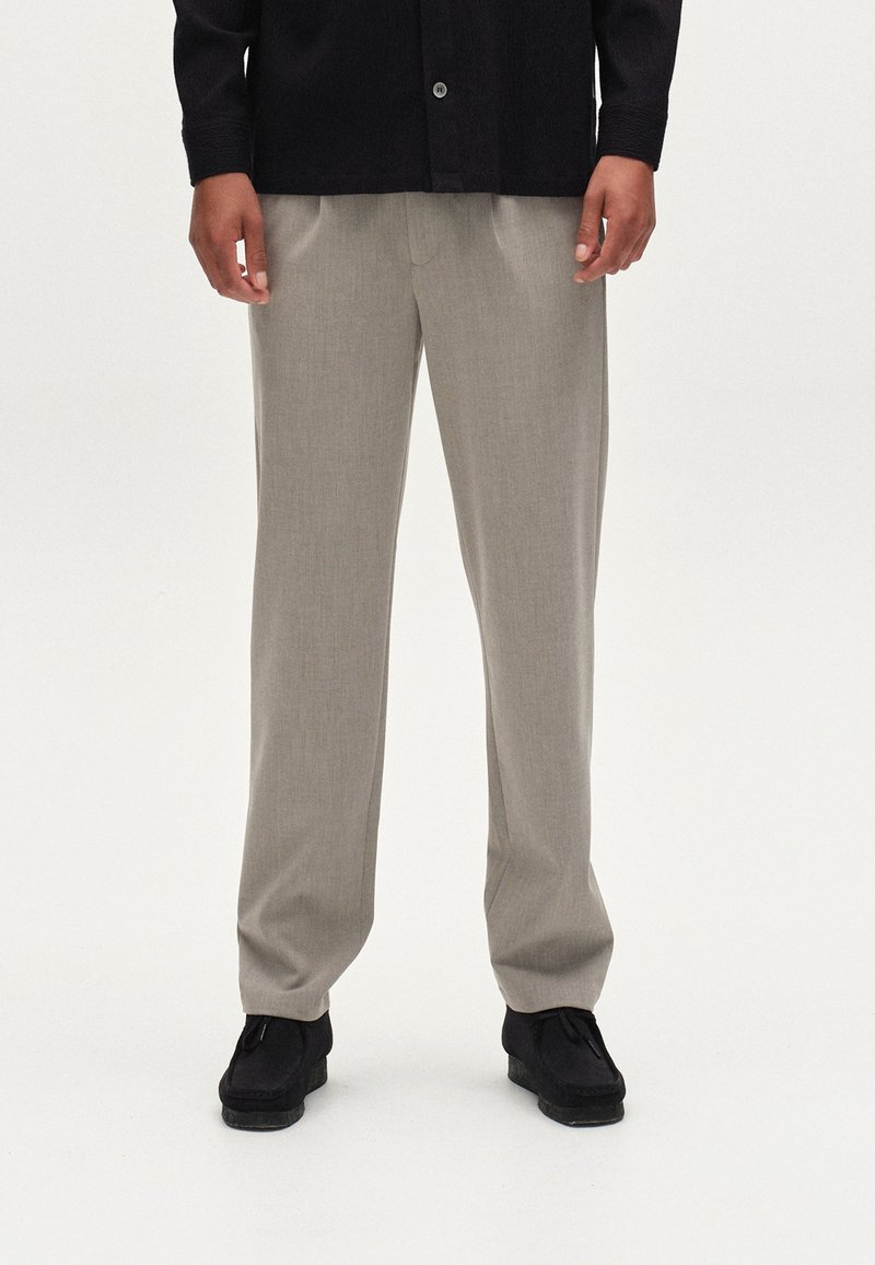 Beige, taillierte Hosen mit glatter Textur, die eine leicht schmalere Passform haben, kombiniert mit schwarzen Schuhen, vor einem neutralen Hintergrund.