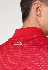 Camisa deportiva roja con una malla texturada y transpirable. Presenta un cuello alzado y un logo impreso en la parte posterior cerca del cuello.