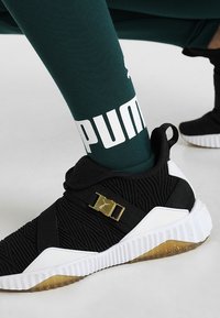 Czarny but sportowy z teksturowanej tkaniny i białą gumową podeszwą. Posiada złote akcenty i czarne paski. Zielona skarpeta z logo "PUMA".