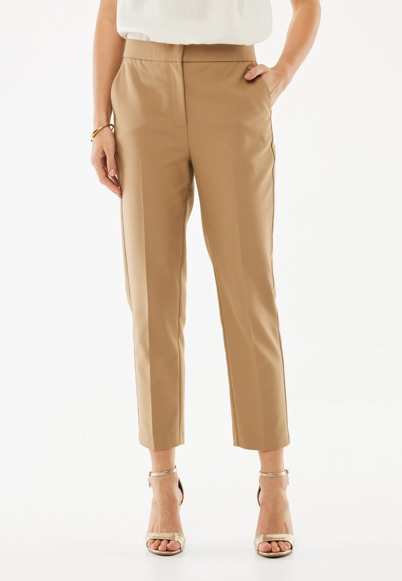 Woman wearing beige tailored ankle pants, white top, gold bracelet, and metallic high-heeled sandals standing with one hand in pocket.