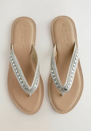 Silverfärgade sandaletter med en pärlbroderad rem, som har en platt tan sål och en strukturerad fotbädd. Sömnadsdetaljer längs kanterna.