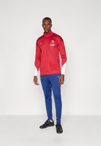 Red athletic jacket with white accents and logo, paired with blue pants and black sports shoes. Smooth fabric and fitted design.