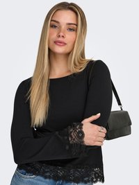 Jonge vrouw met lang blond haar die een zwart shirt met lange mouwen met kanten manchetten en zoom draagt, blauwe spijkerbroek en een zwarte schoudertas.