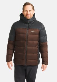 Uomo che indossa un piumino nero e marrone con cappuccio, in piedi e sorridente contro uno sfondo chiaro e semplice.