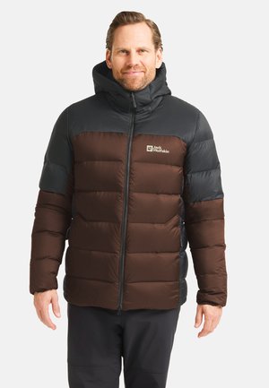 Mann in einer schwarzen und braunen Daunenjacke mit Kapuze, der steht und lächelt vor einem schlichten hellen Hintergrund.