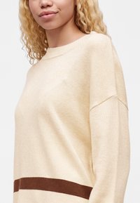 Jeune femme aux cheveux blonds bouclés portant un pull crème avec une rayure horizontale marron près de l'ourlet.
