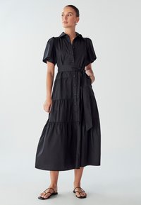 Juodas marškinėlių suknelė iš lengvos medžiagos, su trumpomis pūkuotomis rankovėmis, sluoksniuotu dizainu ir surišta juosmens linija, užtikrinančia prigludusį siluetą.