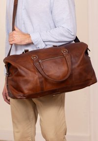 Ruskea nahkainen matkakassi, jossa on kaksi kahvaa, irrotettava olkahihna ja vetoketjullinen etutasku. Sileä pinta, suuri koko.