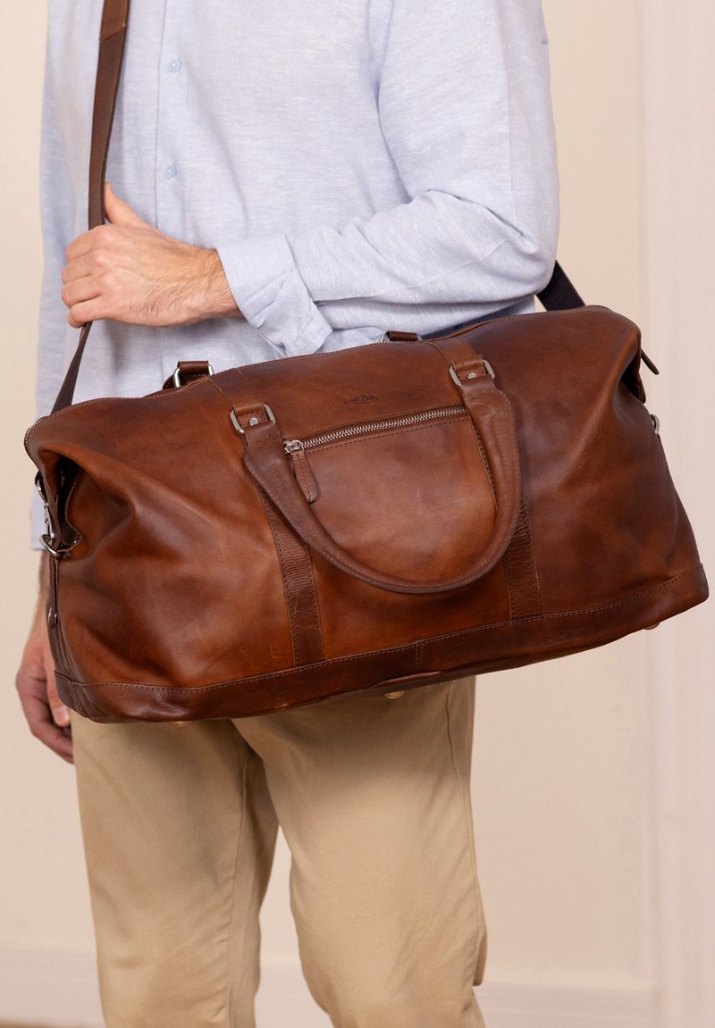 Ruskea nahkainen matkakassi, jossa on kaksi kahvaa, irrotettava olkahihna ja vetoketjullinen etutasku. Sileä pinta, suuri koko.