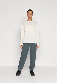 Žena stojaca čelom dopredu v voľnej bielej blúzke s dlhým rukávom, sivých jogger nohaviciach a čiernych teniskách na jednofarebnom pozadí.