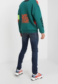 Teal tröja med färgglada mönster och text, i kombination med mörka skinny jeans och chunky vita sneakers med röda och blå accenter.