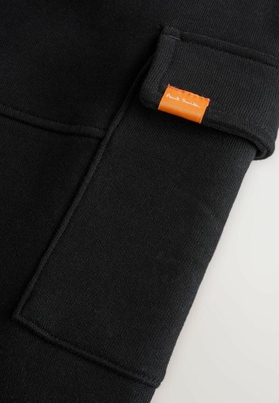 Musta collegepaita, jossa on sileä rakenne, ja ranteessa on näkyvä oranssi etiketti ommeltuna. Siisti muotoilu ja hihansuut ovat resorit.