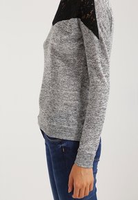 Pull en tricot gris avec un empiècement en dentelle noire sur l'épaule, manches longues et coupe décontractée, associé à un pantalon en denim bleu.