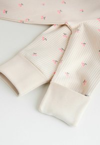 Pehmeä kermainen ribbineulos, jossa on pieniä vaaleanpunaisia kirsikkakuviota ja kiinteät kermansävyiset ribbineulotut hihansuut tai lahkeensuut, aseteltuna tasaisesti valkoiselle pinnalle.