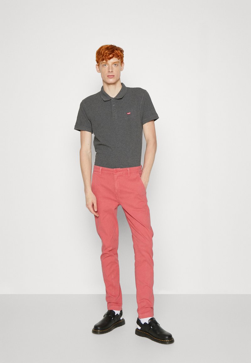Polo gris de corte ajustado, combinado con pantalones rosa ajustados y zapatos negros sin cordones. El polo tiene un pequeño logo rojo en el pecho.