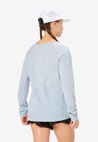 Světle modrá košile s dlouhým rukávem z texturované látky, s kulatým výstřihem a raglánovými rukávy, v kombinaci s černými šortkami a bílou perforovanou čepicí.