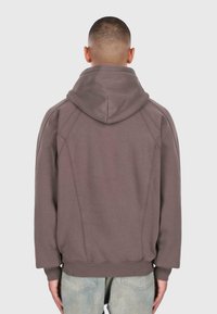 Mężczyzna z krótkimi włosami, ubrany w brązową bluzę z kapturem i jasnoniebieskie dżinsy, pokazany od tyłu na jednolitym jasnym tle.