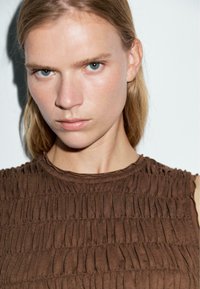 Top marrone senza maniche con pieghe verticali, collo tondo e tessuto morbido e testurizzato. Il design presenta leggere arricciature lungo il collo.
