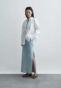 White, long-sleeved shirt, light blue denim skirt with side slits, and brown loafers. Smooth fabric, relaxed fit, minimalistic design.