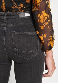 Jeans de mezclilla negros con bolsillos traseros. La tela tiene una textura suave. Una etiqueta en la cinturilla dice "ONLY". Detrás, se puede ver una blusa floral.