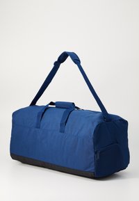Duffel bag i mørkeblå stof med en sort bund. Har to stropper, et håndtag på toppen og en lynlåslomme i siden med mesh-detaljer.