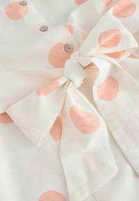 Tissu blanc avec de grands pois roses doux, boutons gris et un nœud noué au centre.