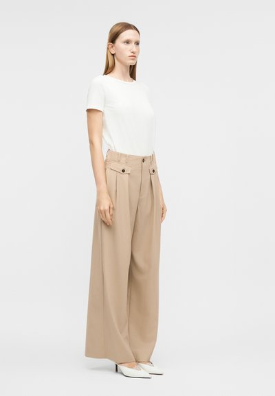 Γυναίκα όρθια, φοράει λευκό κοντομάνικο τοπ, μπεζ παντελόνι ψηλόμεσο με φαρδιά μπατζάκια και κουμπωτές τσέπες, και λευκά μυτερά τακούνια.