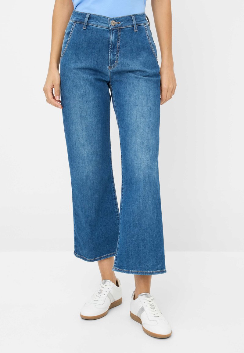 Blauwe denim wijde pijpen jeans met een hoge taille, voorzien van een sluiting met een knoop aan de voorkant, riemlussen en contrasterende stiksels, gecombineerd met witte sneakers.