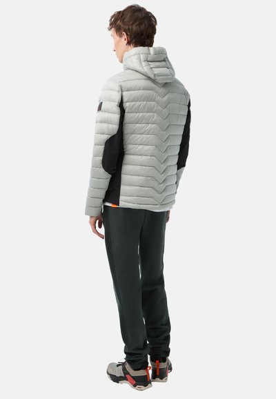 Hellgraue Pufferjacke mit einem Chevron-Steppdesign, schwarzen Ärmeln und Kapuze. Kombiniert mit dunkelgrünen Hosen und mehrfarbigen Sneakern.