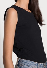 Černý tílko bez rukávů vyrobené z měkké pružné látky. Zaoblený výstřih, přiléhavý střih a boční rozparky. Kombinováno s kraťasy z džínoviny s vysokým pasem.