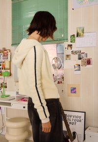 Kremna, puhasta skrajšana kapuca s temno modrimi črtami ob rokavih, v kombinaciji s temnimi kavbojkami. Prostor ima pisalno mizo, zeleno svetilko in stensko dekoracijo.