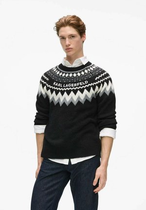 Joven con un jersey de Karl Lagerfeld en negro y blanco con estampado sobre una camisa blanca, combinado con vaqueros azul oscuro, sobre un fondo claro liso.