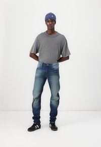 Grå oversized t-shirt, blå denimjeans med blekning, och svarta skor med vita ränder. Man bär en blå ribbad beanie och står.