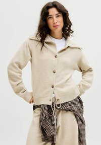 Donna con capelli scuri e mossi indossa un cardigan beige con bottoni, una maglietta bianca e una camicia a quadri legata in vita, in piedi con le mani sui fianchi.