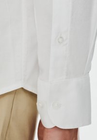 Biała koszula z rękawem z guzikami, z dwoma guzikami, wykonana z fakturowanej tkaniny, w połączeniu z jasnobeżowymi spodniami.