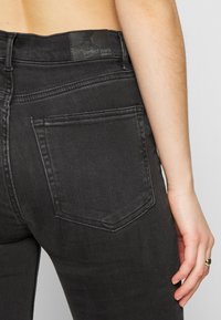 Jeans de denim preto com um design ajustado, apresentando um bolso traseiro limpo e uma etiqueta na cintura com "perfect jeans" gravado em preto.