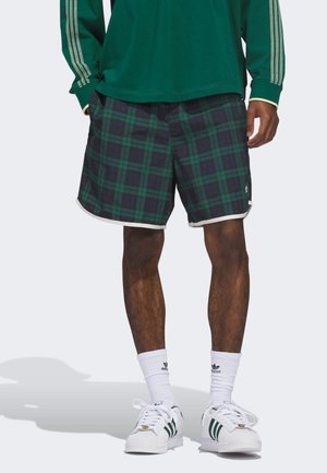 Men's plaid shorts in dark green and navy with white trim. Made from soft fabric featuring a checkered pattern. Paired with white sneakers.