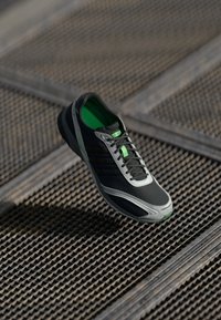 Svarte og grå atletiske sko med mesh-overdel, grønne accenter, snører og gummisåle, vist mot en teksturert bakgrunn.