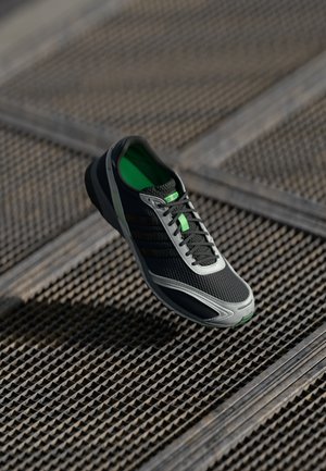 Black and gray athletic shoe with mesh upper, green accents, laces, and rubber sole, displayed against a textured background.