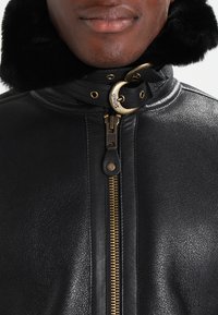 Chaqueta de cuero negro con un cuello de piel sintética, que presenta un destacado zipper dorado y acentos de hardware metálico. Textura suave y diseño clásico.