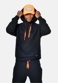 Haut à capuche noir avec des cordons de serrage et des accents orange, doté d'une poche kangourou. Associé à un pantalon de jogging noir assorti et une casquette orange vif.