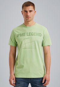 T-shirt en coton vert avec des manches courtes, un col rond et un texte imprimé en vert foncé. Le tissu est lisse et décontracté.