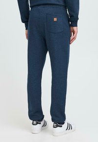Hombre vistiendo pantalones de chándal azul marino con bolsillo trasero y zapatillas Adidas blancas con rayas negras, de pie frente a un fondo liso.