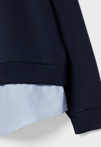 Felpa navy a strati sopra una camicia a righe blu chiaro e bianco, con orlo e polsini a coste, texture del tessuto liscia.