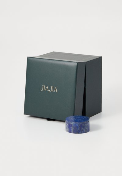 Grön fyrkantig låda med gyllene bokstäver "JIA JIA" och en rund blå sten bredvid. Lådan har en slät yta och ett viklock.