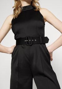 Mono negro con cuello alto y cuerpo ajustado, combinado con un cinturón negro ancho que tiene una hebilla rectangular y acentos de hardware plateado.