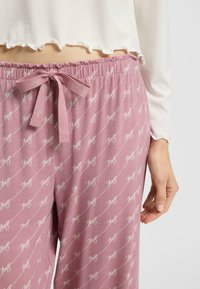 Roze pyjamabroek met een herhalend wit logopatroon, elastische tailleband met een strik aan de voorkant en een relaxed fit. Zachte stofstructuur is zichtbaar.