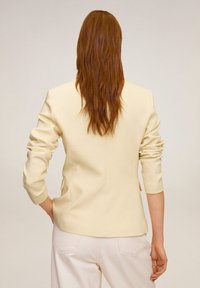 Blazer su misura color crema con design aderente, maniche lunghe e morbidi spalline. Texture liscia, dettagli minimalisti e spacco posteriore.
